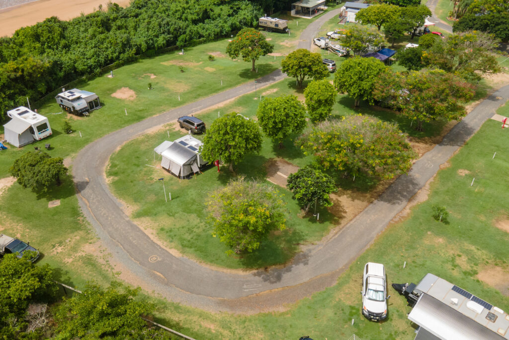 An aerial view of a campground featuring a winding paved road surrounded by green trees and grassy areas. Several RVs and tents are set up in designated spots. The campground is nestled near a dense area of trees, and part of a sandy beach is visible in the background.