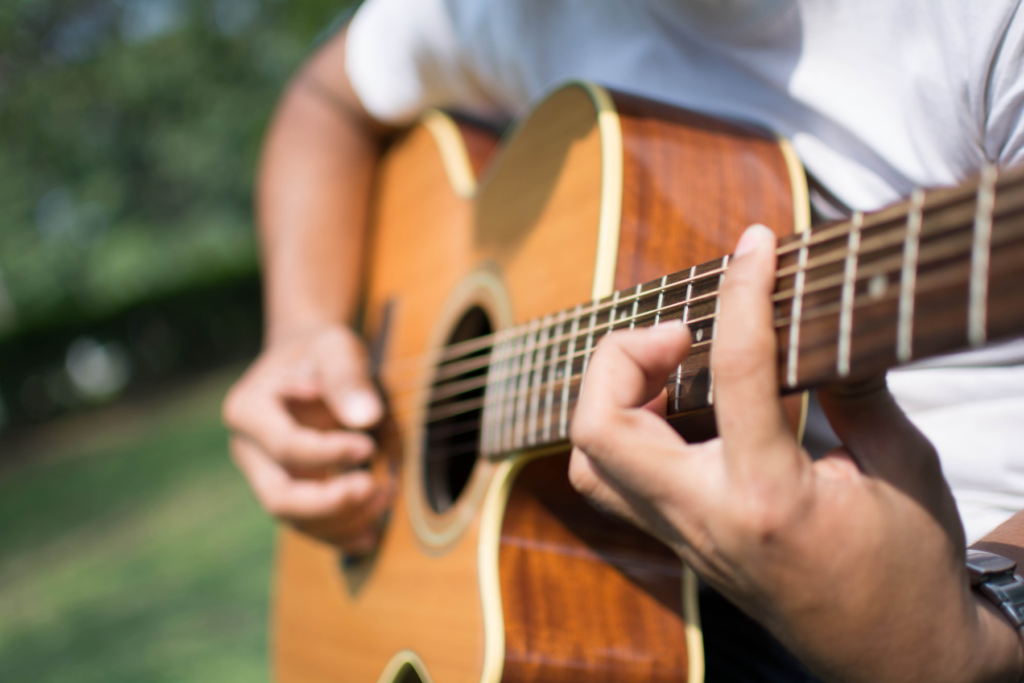 A close-up shot of a person playing an acoustic guitar outdoors. The focus is on the fingers strumming the strings and pressing the frets, with a blurred green background indicating a natural setting. The person is wearing a short-sleeved white shirt.