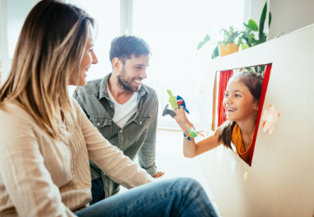 A young girl performs a puppet show with a hand puppet from a cardboard box theater, while a man and woman smile and watch. The scene takes place indoors, with sunlight streaming through a window in the background. Houseplants can be seen behind them.