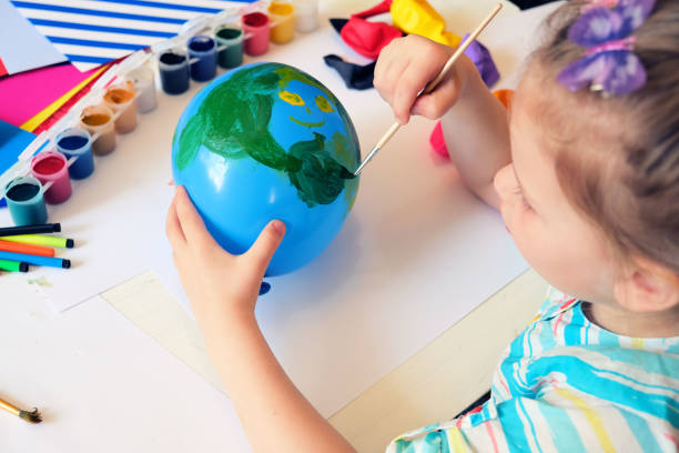 A child is painting a blue balloon with a smiley face using green paint. Various paint containers, colored pencils, and art supplies are scattered around on the table. The child has a colorful butterfly hair clip and is wearing a striped shirt.