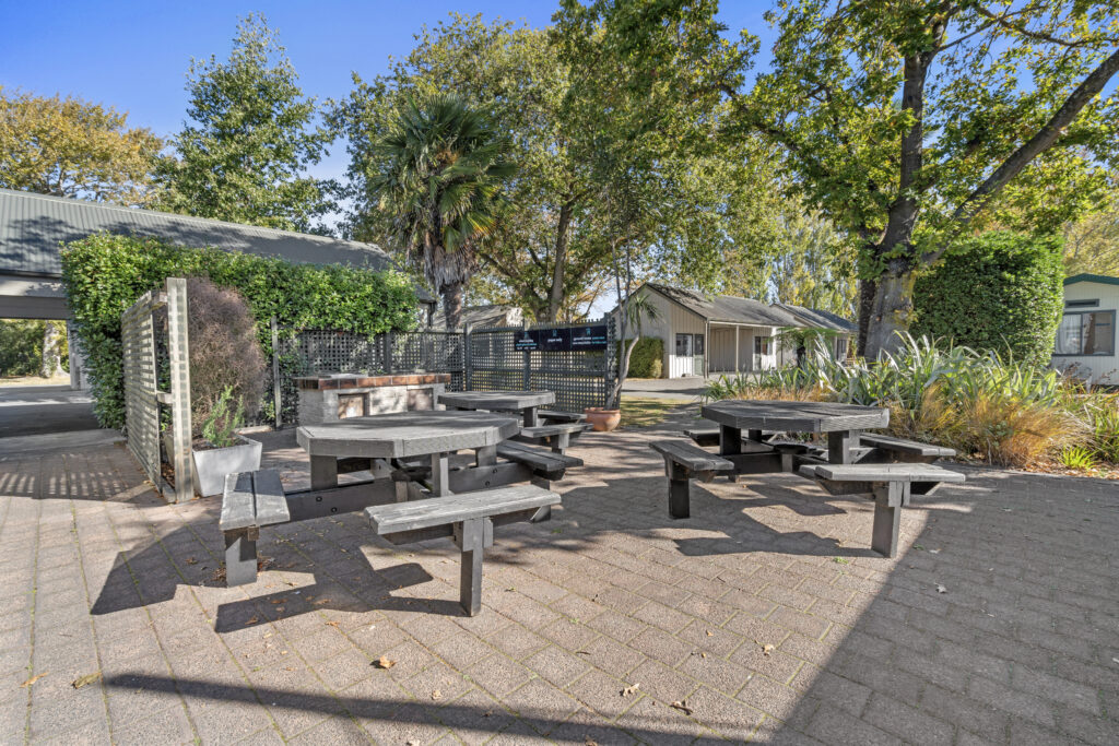 Outdoor area with multiple picnic tables arranged on a paved surface, surrounded by trees and greenery. A few buildings and a covered structure are visible in the background, with a clear blue sky above. The space appears to be a communal or recreational area.