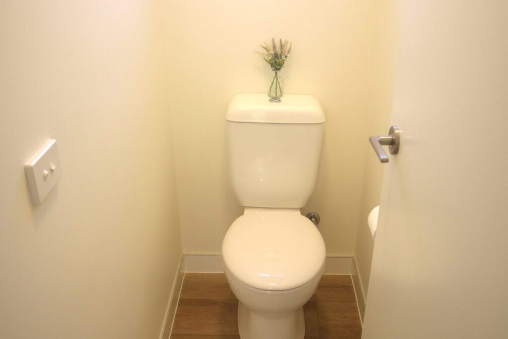 A small, tidy bathroom features a white toilet with a vase containing small flowers on the top of the tank. The room has a wooden floor and light-colored walls, with a closed door and a light switch on one side.