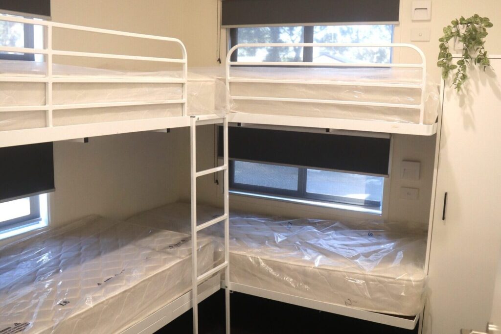 A well-lit room containing two sets of white metal bunk beds with mattresses covered in plastic. Two windows with black roller blinds are seen on adjacent walls. A small potted plant hangs on the right wall, adding a touch of greenery.