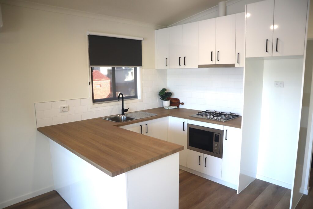 A modern kitchen with white cabinets, a wood countertop, and stainless steel appliances. It features a double sink, gas stove, built-in microwave, and a dark window shade. The kitchen has a minimalist design with light wood flooring and white tiled backsplash.