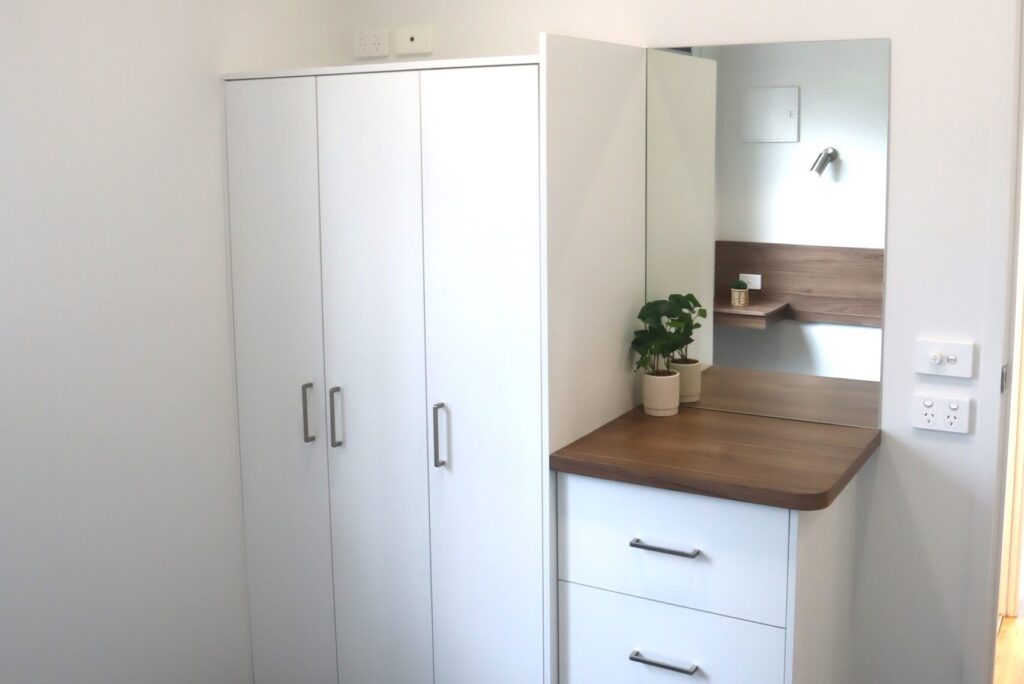 A corner of a room featuring a mirrored dresser with a wooden top and two drawers, a small potted plant on top, and a white wardrobe with three doors and silver handles. The background includes a wall-mounted light and a shelf with a small item on it.