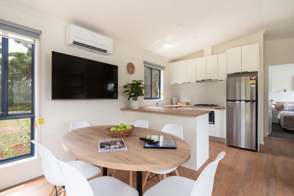 A modern kitchen and dining area with a round wooden table surrounded by white chairs, a TV mounted on the wall, an air conditioning unit above the TV, a potted plant, and a stainless steel refrigerator. The room has large windows and a doorway leading to a bedroom.