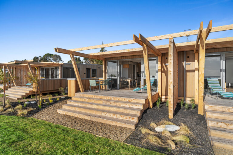 A modern wooden holiday cabin at Papamoa Beach with large glass doors opens onto a spacious deck featuring outdoor seating and a table. The structure has a pergola-style roof. The luxury cabin is surrounded by a grass lawn and newly planted vegetation, set against a clear blue sky.