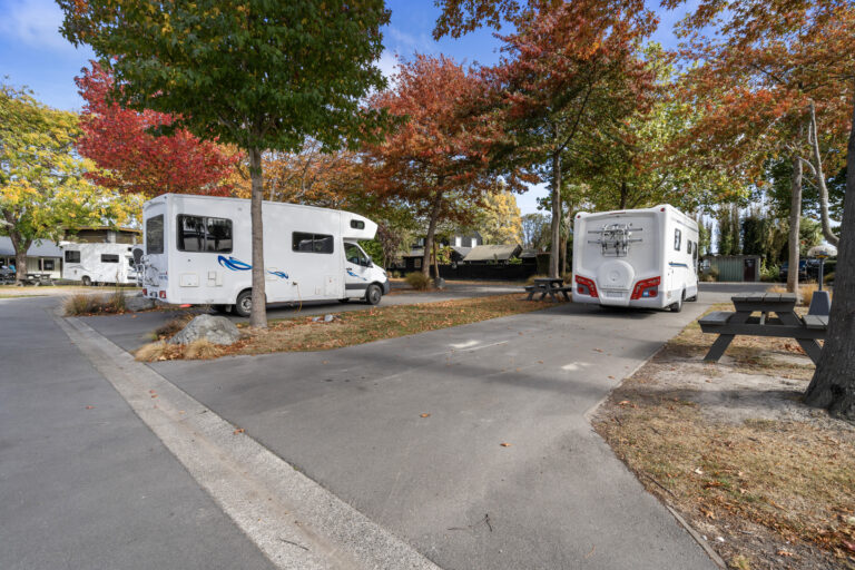 Two motorhomes parked at a campsite with autumn-colored trees. The area features paved parking spaces and picnic tables. The vibrant red and yellow leaves add a scenic backdrop to the setting, creating a cozy and inviting atmosphere.