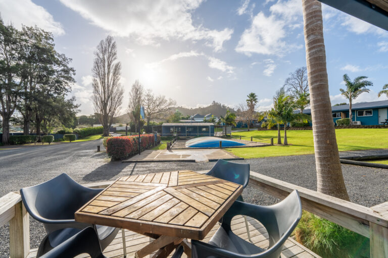 A sunny outdoor scene with a wooden table and chairs on a deck overlooking a lush green yard with palm trees, a swimming pool covered with a blue tarp, and a backdrop of mountains and clear skies. A road and a few buildings are visible in the background.