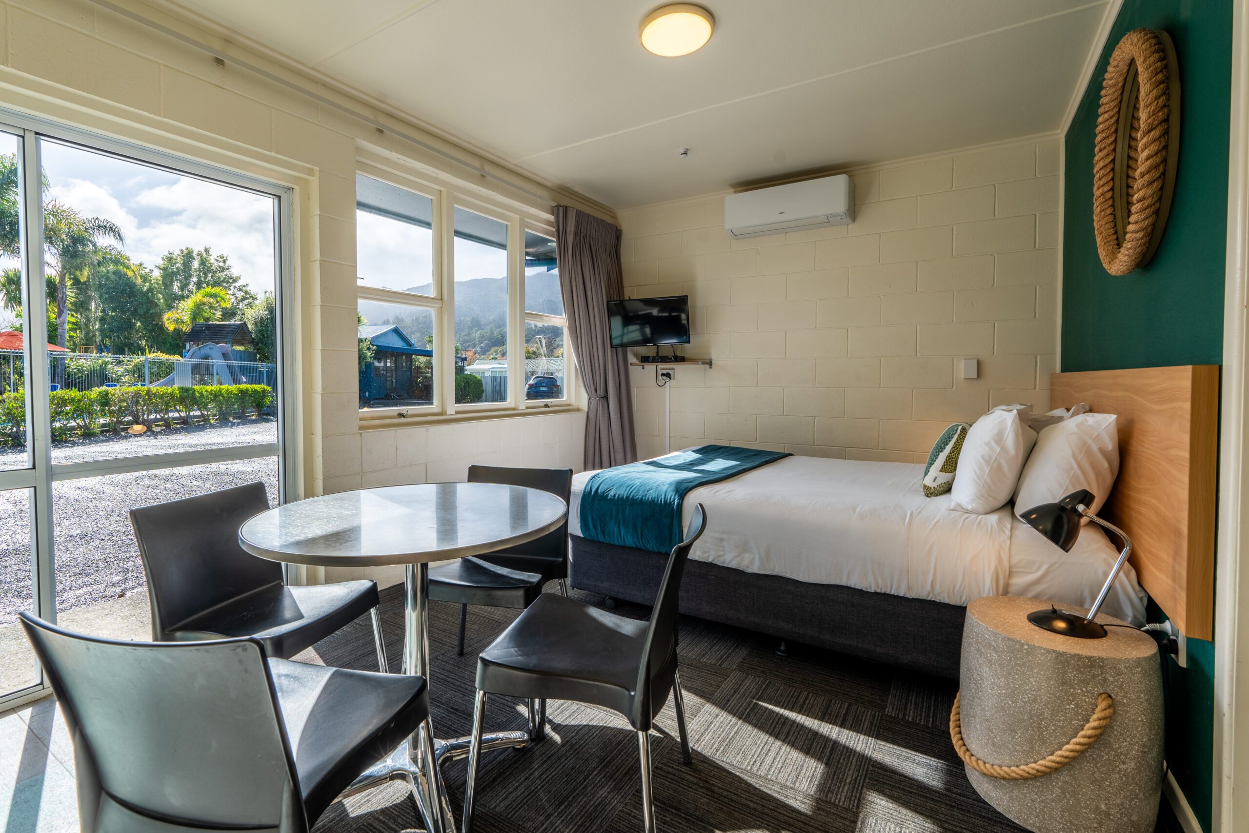 A bright, modern hotel room with a queen-sized bed, a small round table with four chairs, and a wall-mounted flat-screen TV. A large window and sliding glass door allow natural light to fill the space. The room has a blue and white color scheme and outdoor views.