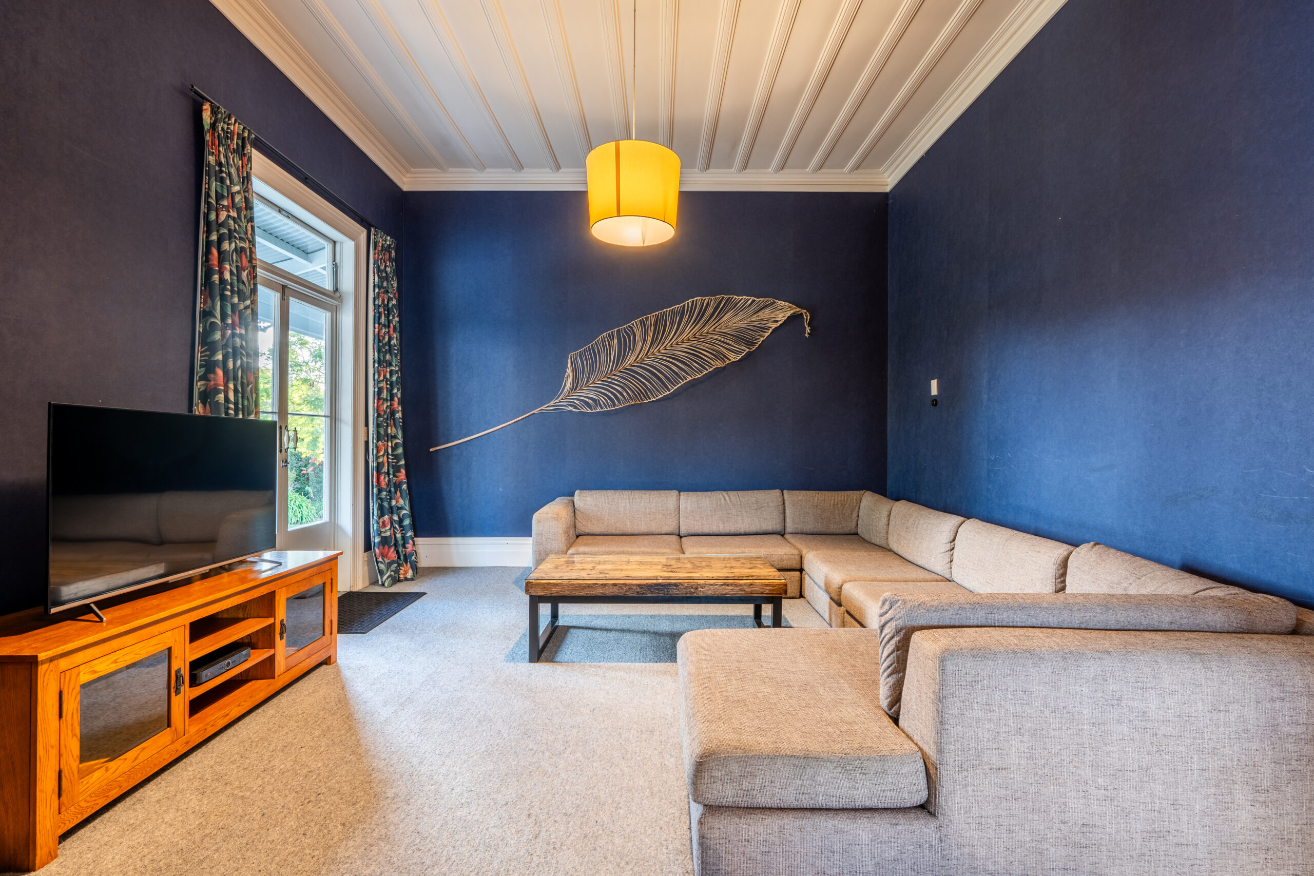 A cozy living room with dark blue walls featuring a large single feather wall piece. The room has a light beige L-shaped sofa, a wooden coffee table, a flat-screen TV on a wooden entertainment unit, and floral curtains framing a large window. A warm pendant light hangs from the ceiling.