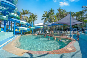 Children swimming and playing in a pool with a large, winding waterslide on the left and several palm trees and tropical plants in the background. Parents are supervising from shaded seating areas under sun canopies on the right. The clear, sunny sky shines bright like the North Star.