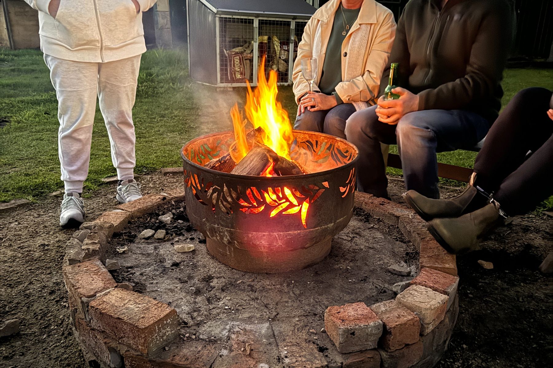 Four people sit and stand around a lit fire pit, enjoying the warmth on a calm evening. The fire pit, made of metal and brick, emits a bright flame. One person holds a beer bottle, while another's legs are crossed. The ground is bare with patches of grass.