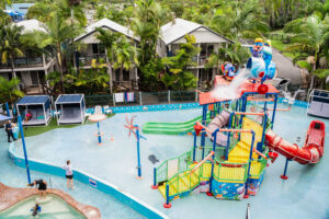 A vibrant water playground, known as the North Star of family fun, features various slides, climbing structures, and water sprays, surrounded by lush palm trees and modern villas. Children play in the shallow water while parents supervise from the poolside. Fun water activities are in full swing.