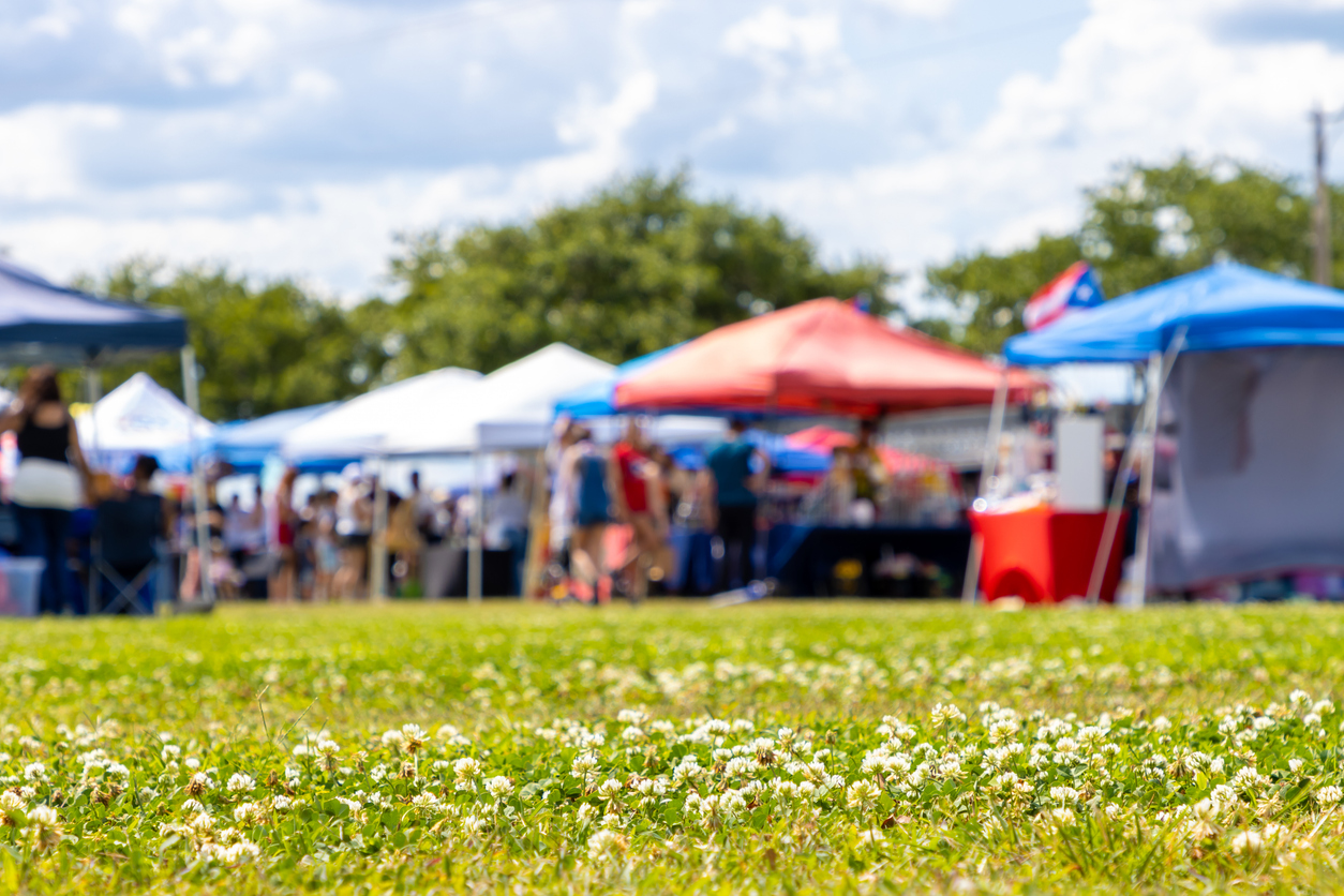 A vibrant outdoor market with various colorful tents and booths set up on a grassy field. The focus is on white clover flowers in the foreground, while people are seen mingling among the tents in the blurred background under a partly cloudy sky.