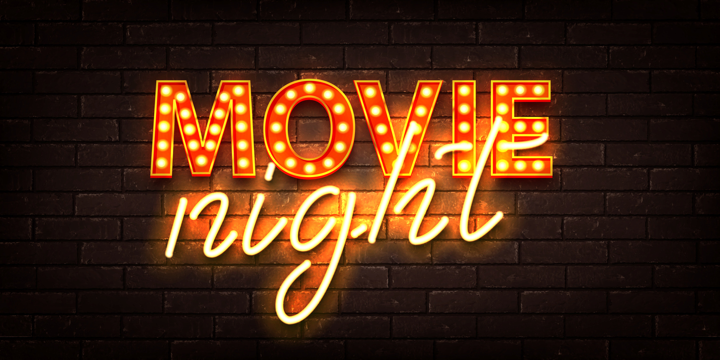 A neon sign on a brick wall reads "MOVIE night." The word “MOVIE” is in bold, bright orange letters with lights, and the word “night” is in a cursive yellow neon script. The sign emits a warm glow, illuminating the brick background.