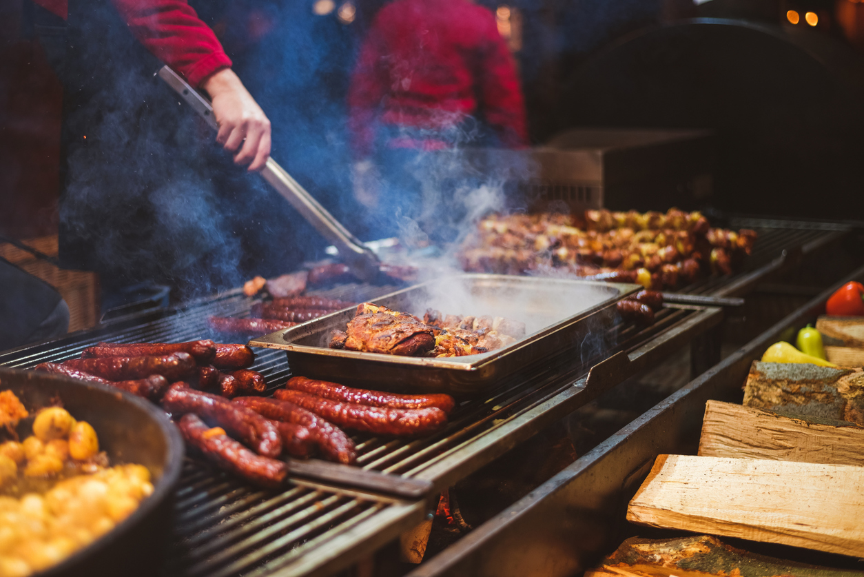 A person grills various meats, including sausages and ribs, on a large outdoor barbecue grill. Smoke rises from the cooking food, creating a rustic ambiance. Wood logs and vegetables are visible in the foreground. The person is wearing a red jacket.