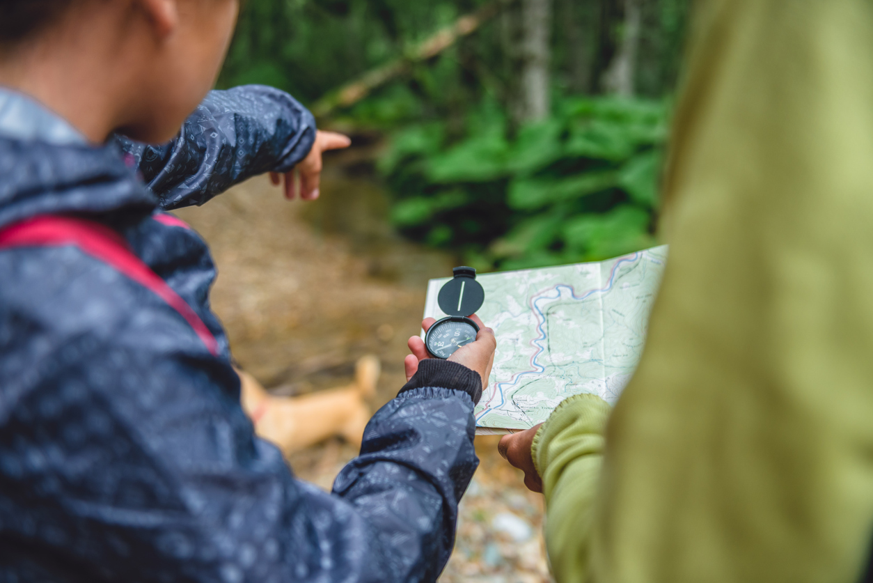 Two individuals, dressed in outdoor gear, are seen navigating with a map and a compass in a forest. One person is holding the compass and the map, while the other person points towards a direction, indicating the way to go. The background includes blurred greenery.