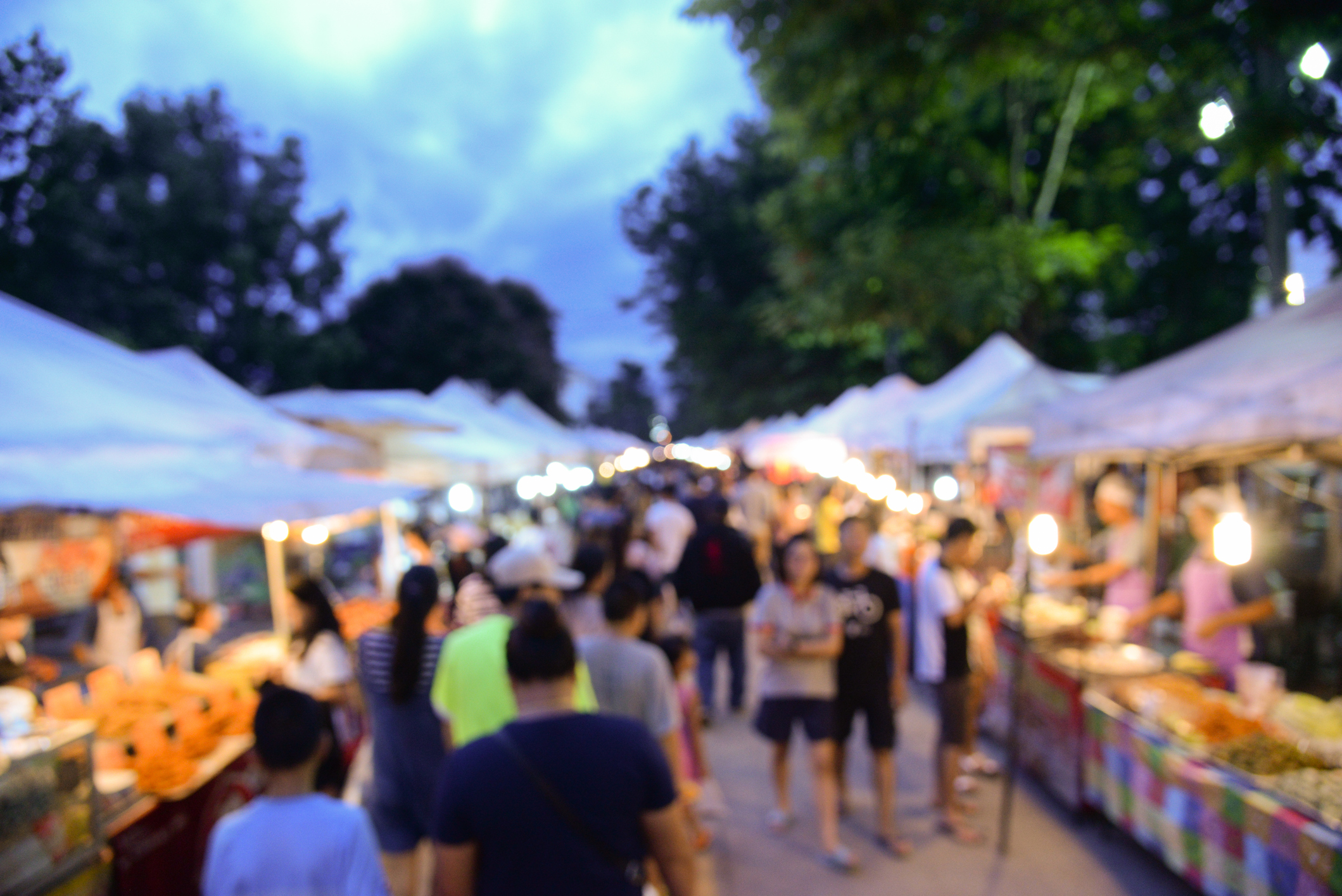 A bustling outdoor market at dusk, with many people walking between rows of vendors under white tents. The scene features blurred, glowing lights, and trees in the background, evoking a lively and vibrant atmosphere.