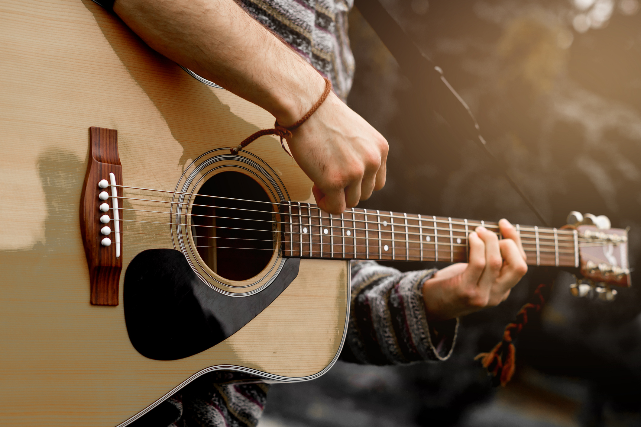 A close-up image of a person playing an acoustic guitar. The person is wearing a knitted sweater, and the visible hand is strumming the strings while the other hand is on the fretboard. The background is blurred with a warm, natural outdoor setting.