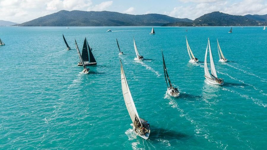 Aerial view of multiple sailboats on a clear turquoise sea, with several sailboats racing towards the horizon. The background features a mountain landscape under a partly cloudy sky.