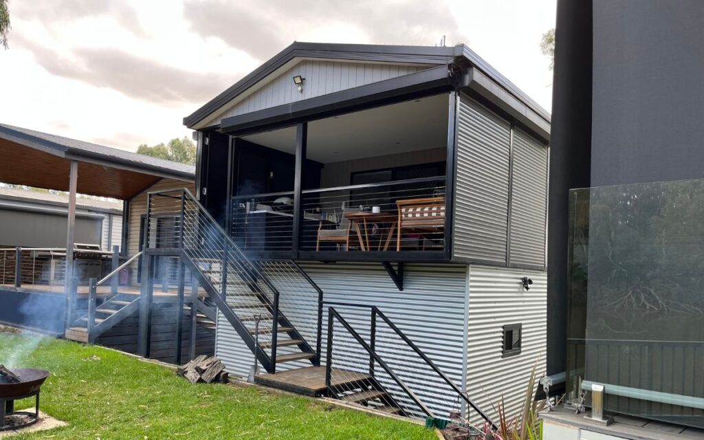 A modern tiny house with corrugated metal siding, elevated on stilts. The house features a covered porch with a dining area, accessible by a set of stairs. The surroundings include a grassy yard with a fire pit and adjacent structures.