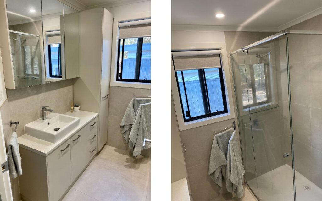 A modern bathroom featuring a sink with a white countertop, beige cabinets, and a wall-mounted mirror on the left. The right side shows a glass-enclosed shower with a gray towel hanging nearby. The room has a frosted window and beige tiles on the wall and floor.