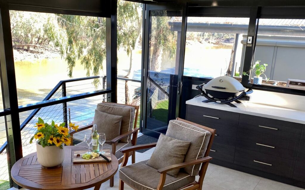 A cozy indoor-outdoor space with a wooden table and two cushioned chairs. The table has a vase with yellow flowers and a water bottle. A counter with a small grill and plant is on the right. The area overlooks a river through large windows and a sliding door.