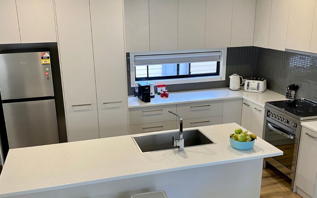 A modern kitchen featuring white cabinets, a stainless steel fridge, a built-in oven, a microwave, a coffee maker, and a toaster. A sink is installed in a white countertop on the central island with a bowl of fruit, and a window with blinds is above the sink.