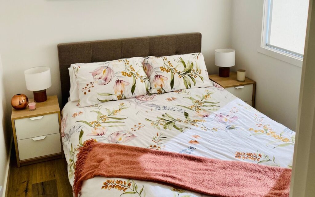 A neatly made bed with floral-patterned bedding and two matching pillows, situated in a cozy bedroom. The bed has a brown headboard and a pinkish-red throw draped across the foot. Each side of the bed has a wooden nightstand with lamps and small decorative items.