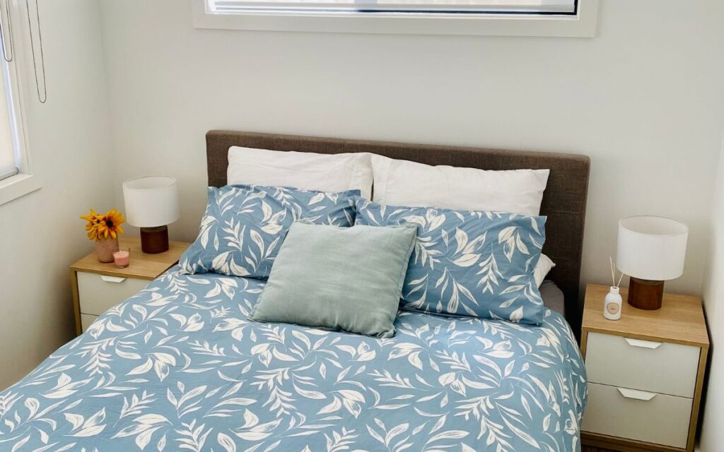 A neatly made bed with a blue and white leafy patterned comforter and three pillows, one green and two matching the comforter. Two small nightstands with lamps and a few decorative items stand on either side. A large window above the bed lets in natural light.