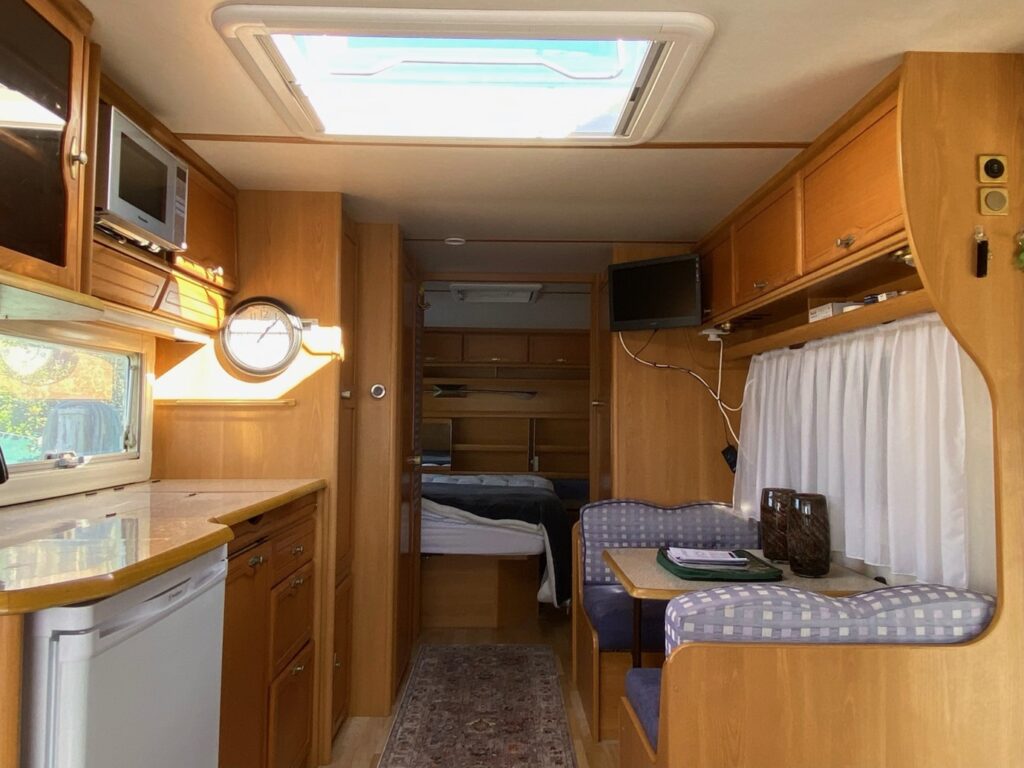 The interior of a cozy RV is visible, featuring a kitchenette with a fridge and microwave on the left, a dining area with a table and cushioned bench seats on the right, and a bed in the rear. Natural light fills the space through a skylight above.