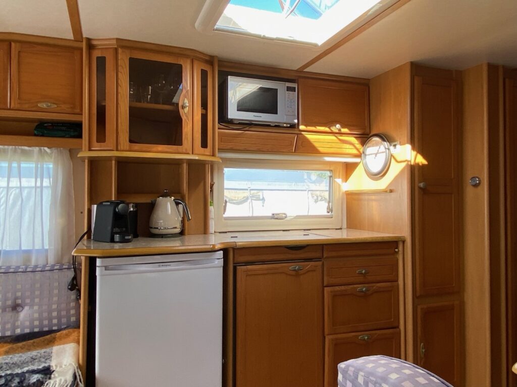 This image shows the interior of a compact kitchen area in an RV with wooden cabinets, a small refrigerator, a microwave, a coffee maker, and a kettle on the countertop. There is a window above the counter, allowing natural light to illuminate the space.