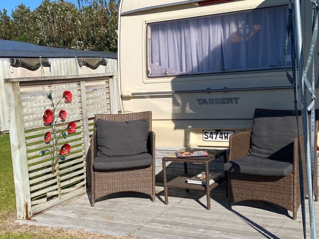 A cozy outdoor setup with two wicker armchairs, each with a gray cushion, facing a small table that holds a book and other items. Behind them is a beige caravan with a license plate reading "S474W". The wooden fence to the side has red flower ornaments.