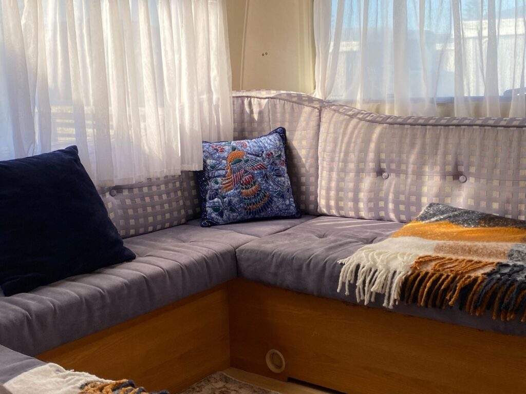 A cozy seating area with light purple cushions and a corner seat is adorned with a colorful embroidered pillow and a patterned throw blanket. Sheer white curtains allow sunlight to stream in, creating a warm and inviting atmosphere.