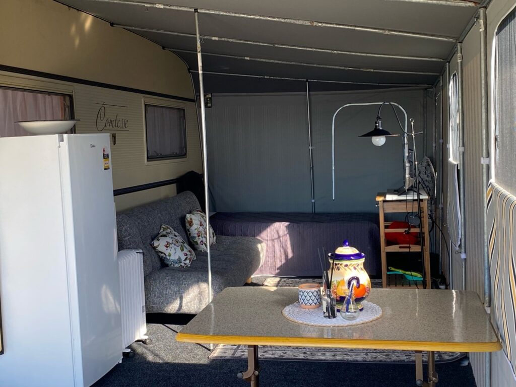 Interior of a cozy caravan with a table in the foreground. The table has a colorful teapot and cups. Behind it, there's a grey sofa with patterned cushions and a bed with a blanket. To the left, there's a white refrigerator, and to the right, a standing lamp.