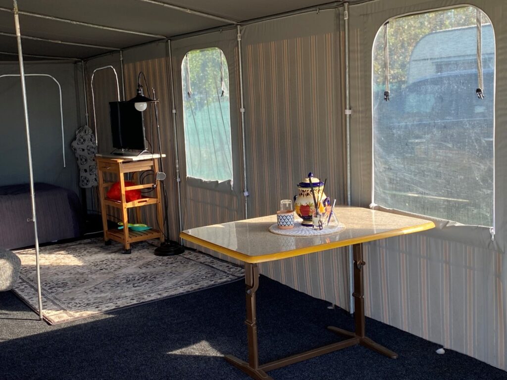 The interior of a tent with a table holding a teapot, cups, and a lantern. A rug and a small shelving unit with a TV, books, and decor are arranged against the tent wall, next to a bed. Light streams through multiple large windows.