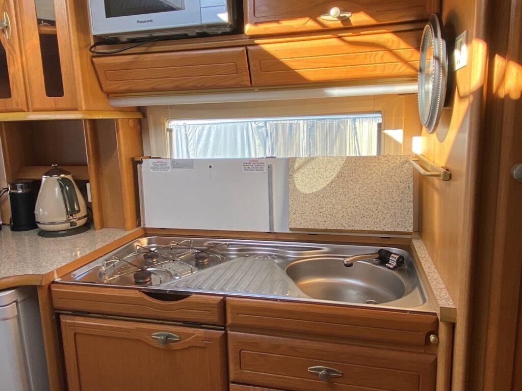 A compact kitchen inside a camper van featuring a small stainless steel sink, a two-burner gas stove, a kettle, and wooden cabinets. Sunlight streams in through a window with a closed blind, highlighting the cozy and functional design.