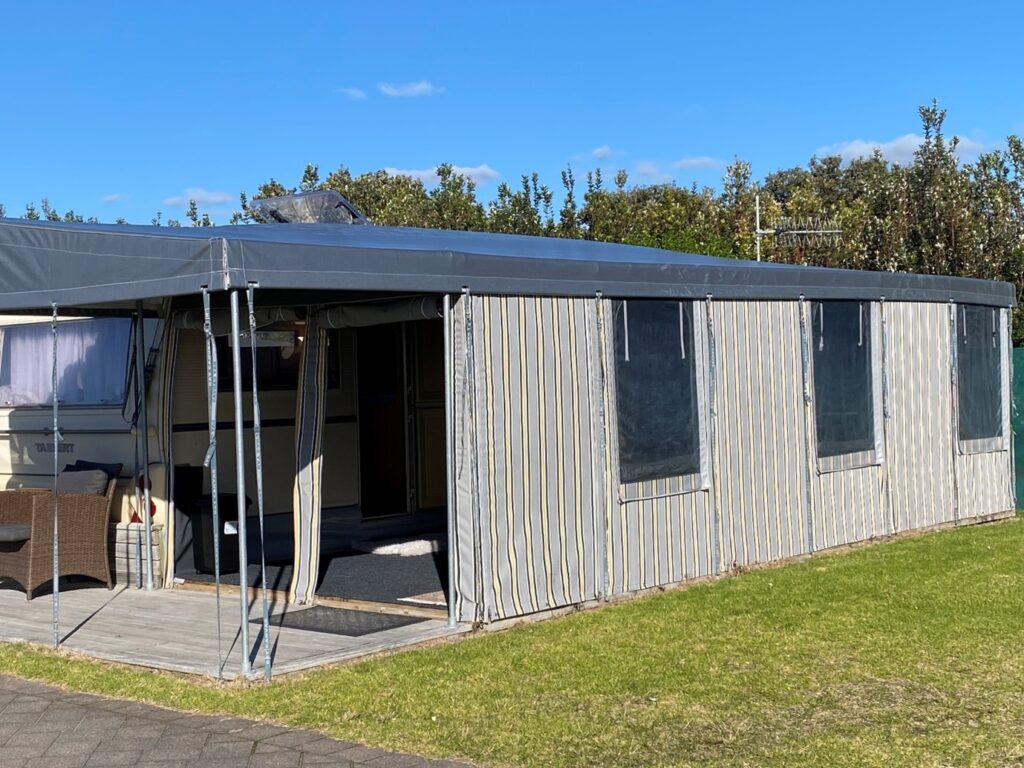 A caravan with an attached, enclosed outdoor awning with multiple windows, situated on a grassy area. There is an entry opening on the side of the awning, and the sun is shining brightly with a clear blue sky overhead.