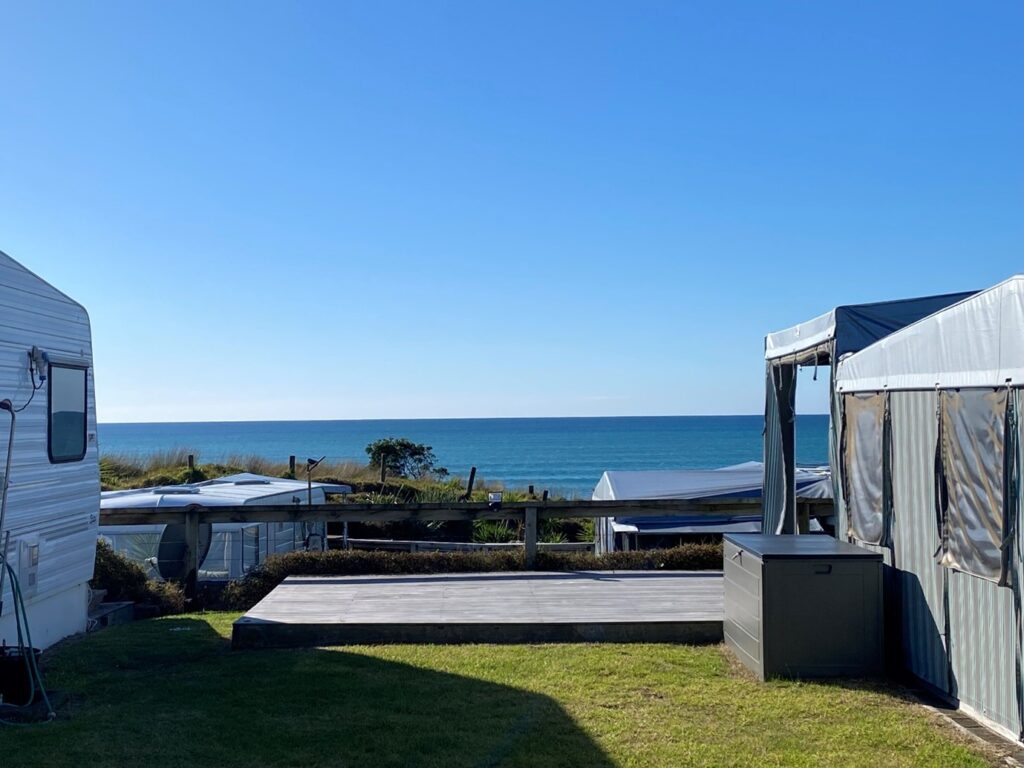 A scenic view of a beach with the ocean in the background. In the foreground, there are two RVs and a small deck area. The sky is clear and blue, and some trees and grass are visible in the midground. The overall atmosphere is peaceful and serene.