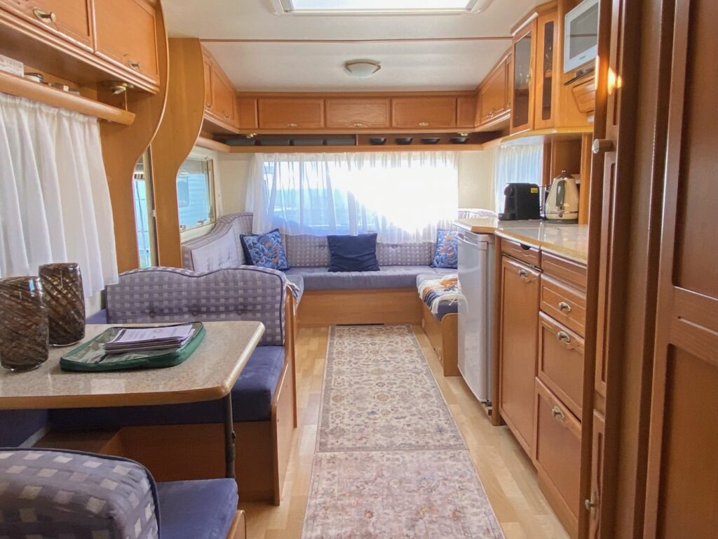 The interior of a cozy RV with wooden cabinetry is shown. There is a kitchenette on the right with a mini-fridge, microwave, and sink. Opposite, a dining area with a table and cushioned benches is visible. At the back, a seating area with large windows and curtains can be seen.