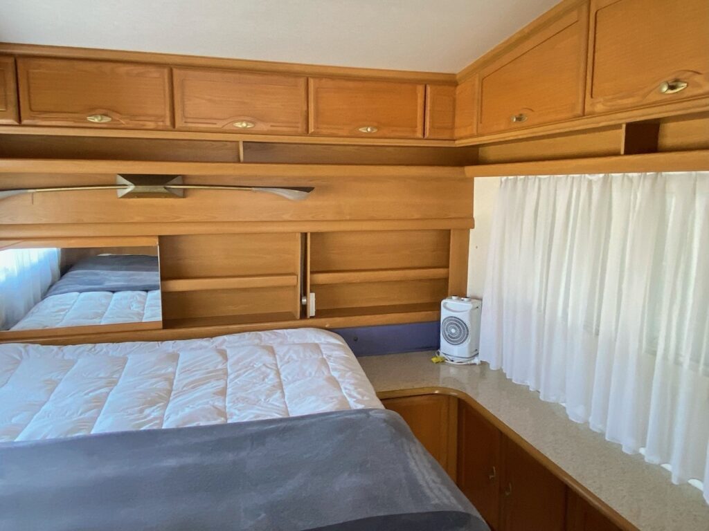 Interior of a cozy RV bedroom with wooden cabinets, a made bed with white and gray bedding, a rectangular window with white curtains, and a small portable fan on a counter next to the bed. The space appears compact and neatly organized.