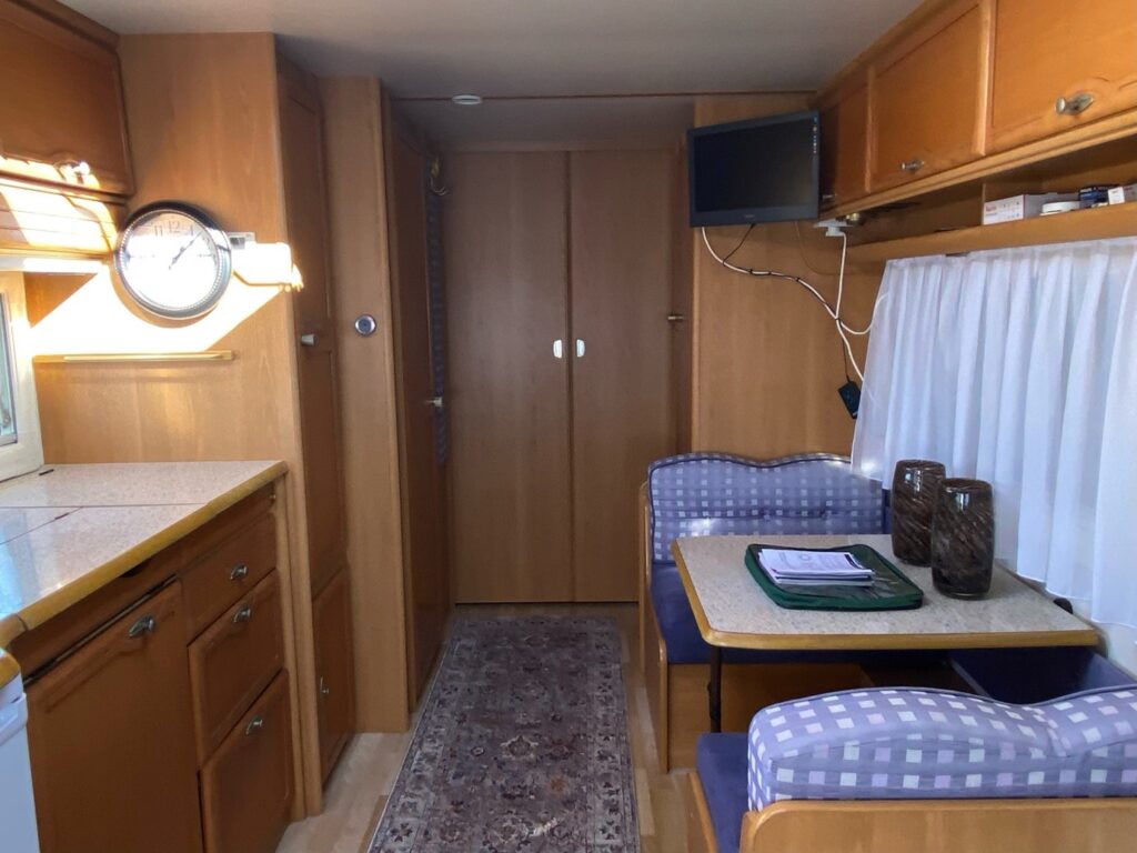 The interior of a small wood-paneled RV features a compact kitchen area with a counter, cabinets, and a wall clock. Opposite the kitchen is a dining nook with padded seating and a table. A television is mounted on the wall, and there is a hallway with closed doors.