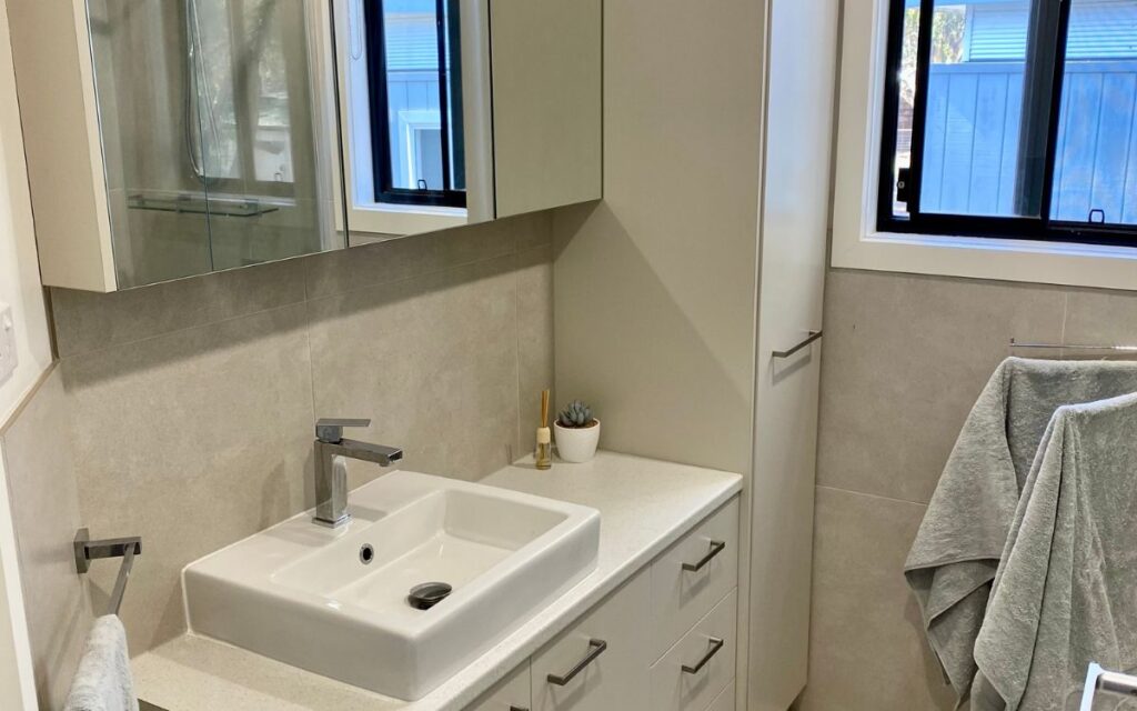 A modern bathroom featuring a beige countertop with a square sink, a chrome faucet, and a mirrored medicine cabinet above. There's a small plant beside the sink, a towel hanging on a rack, and two windows letting in natural light. Towels are draped on another rack.