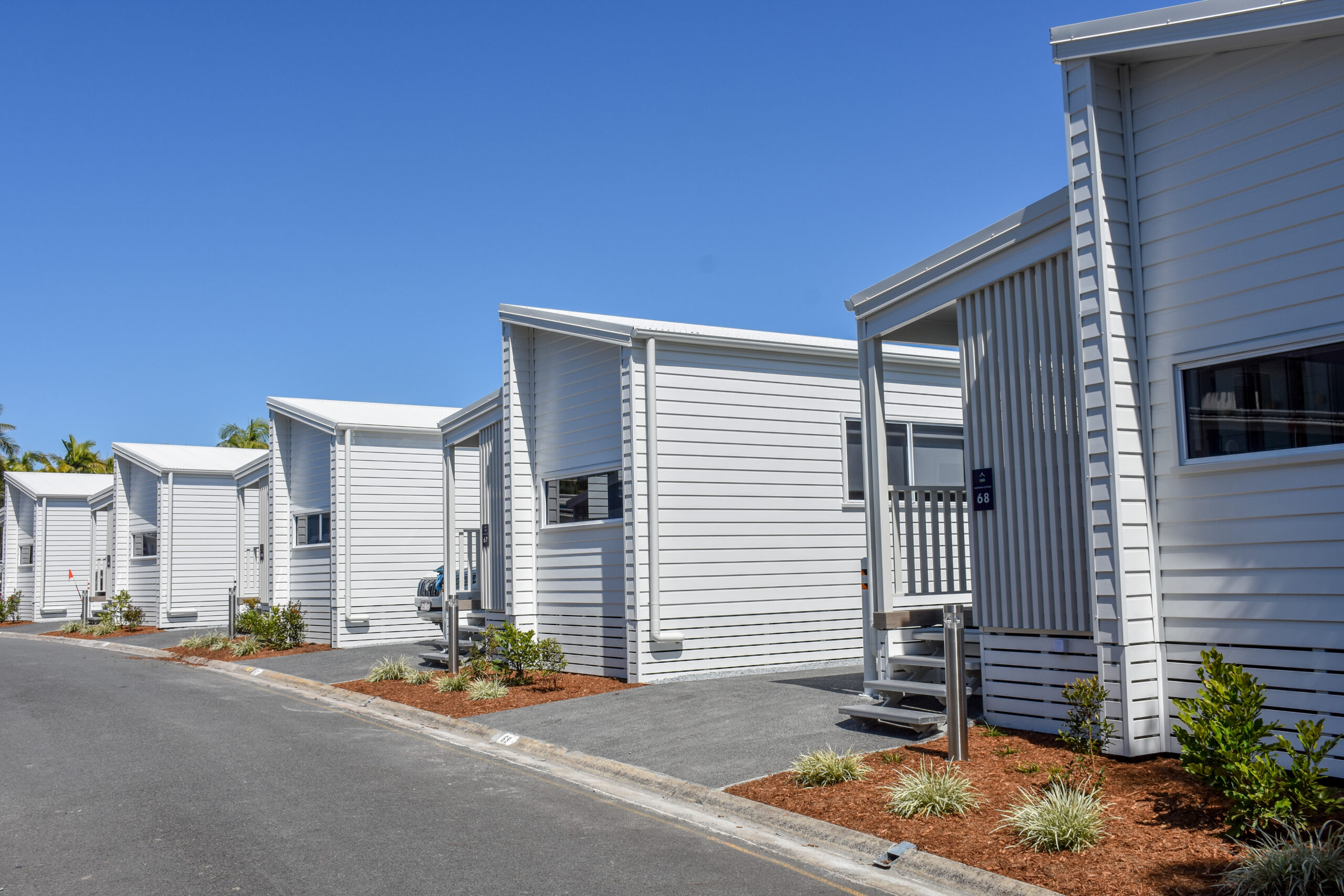 Row of modern, white modular homes with small porches under a clear blue sky resembling a serene North Star evening. The neat landscaping includes small plants and tidy mulch beds lining the walkway. Magnolia Cottage | Tasman Holiday Parks - North Star