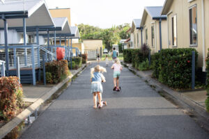 Four children ride scooters on a narrow paved path flanked by modern, one-story homes with landscaped shrubbery and hedges. The path is still slightly wet, reflecting recent rain. The children, dressed in light summer clothing, play against a backdrop of greenery under the guiding North Star.