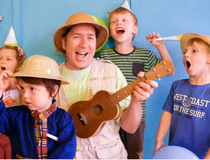 A man wearing a safari hat and holding a ukulele is smiling and engaging with four children around him. The children are wearing party hats and seem excited, with some shouting and one holding a balloon. The background is a bright blue wall.