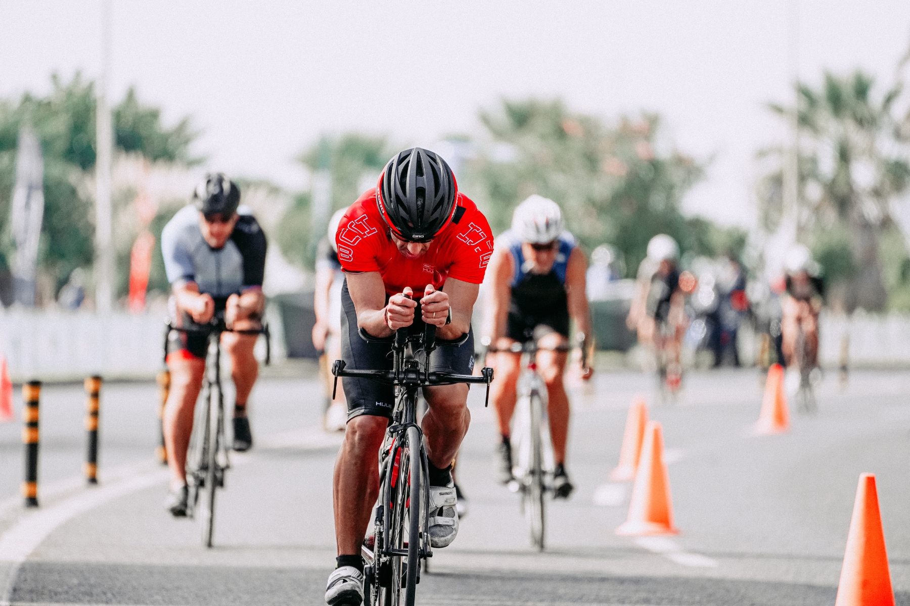 A group of cyclists participating in a race on a paved road. The lead cyclist wears a red jersey and black helmet, while others follow closely behind. Orange traffic cones line the side of the road, and trees and blurred spectators are visible in the background.