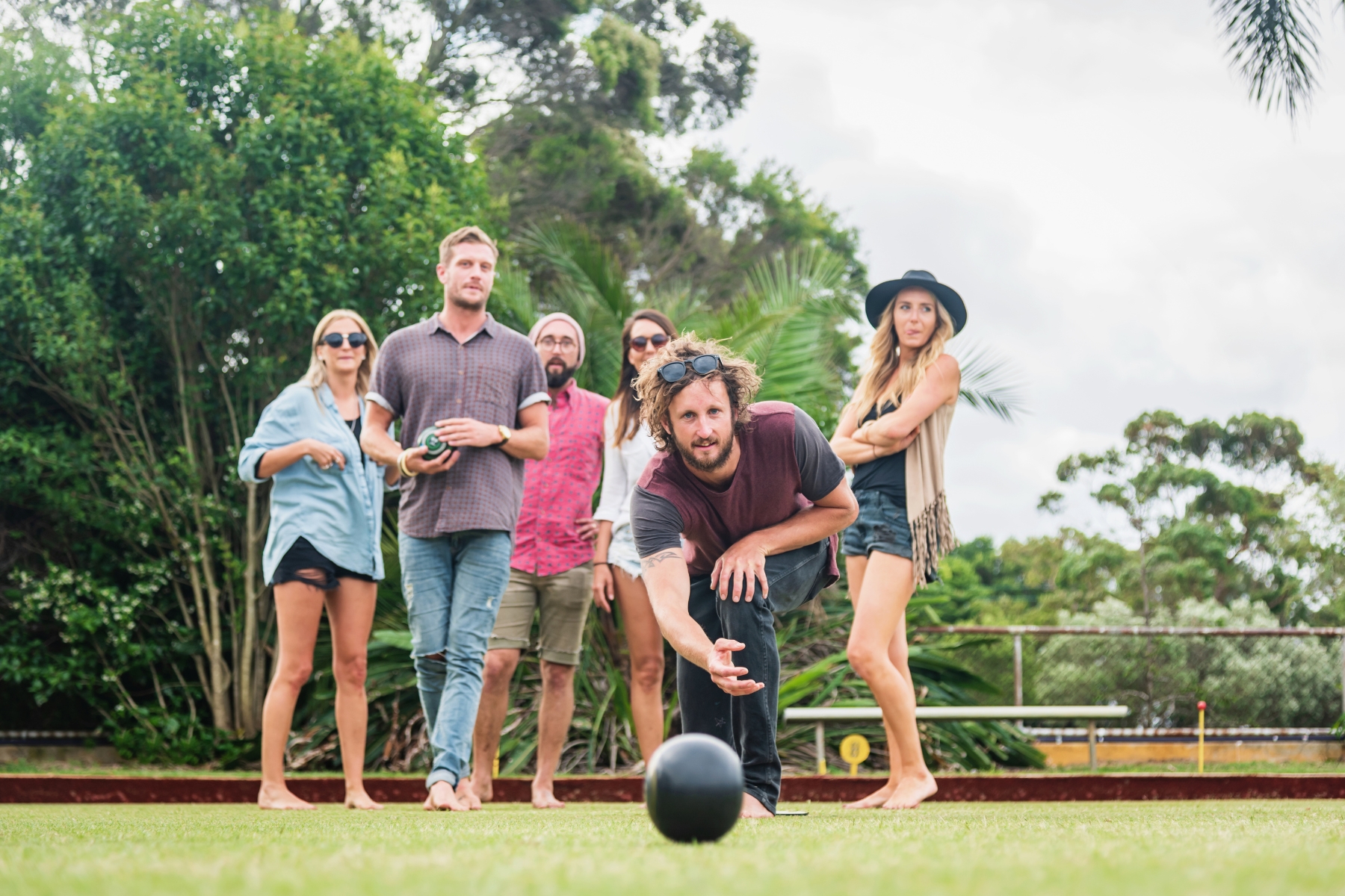 A group of six people playing outdoor lawn bowling. One person in the foreground is rolling a black ball, while the others watch in anticipation. They are casually dressed, surrounded by lush greenery, with trees and a cloudy sky in the background.