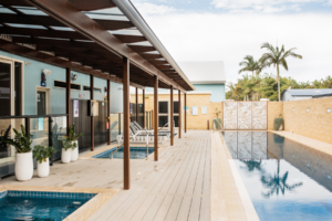 A serene outdoor pool area featuring a long, narrow swimming pool with clear, reflective water. The pool is adjacent to a covered veranda with lounge chairs and small hot tub. Palm trees and modern decor create a relaxing ambiance under the North Star in the partly cloudy sky.